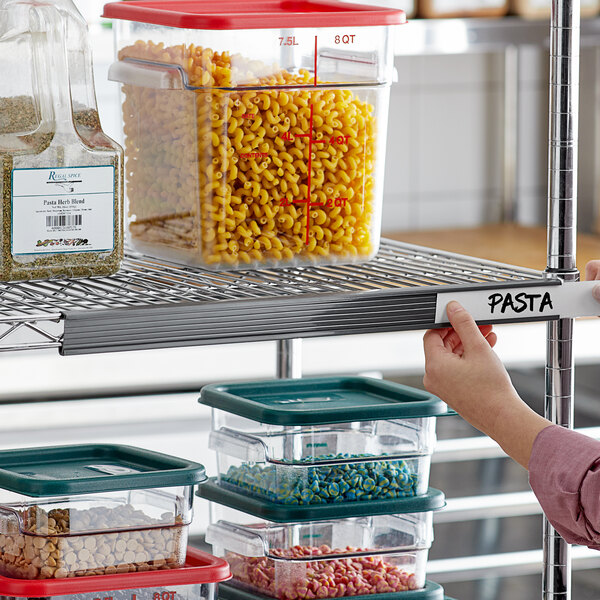 A hand holding a gray plastic label with "pasta" in front of containers of pasta and spices on a shelf.