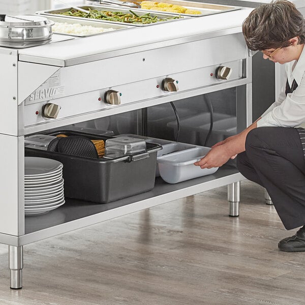 A woman kneeling down in front of a ServIt electric steam table on a counter in a school kitchen.