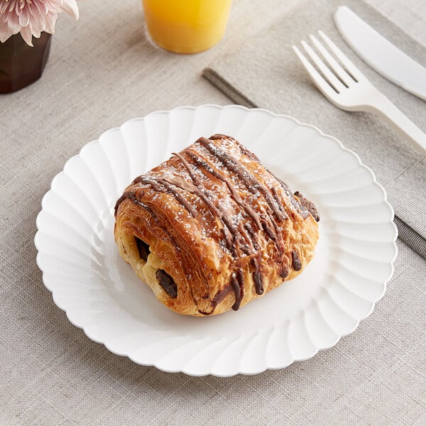 A pastry on a WNA Comet white plastic plate with a fork and knife on a table with a glass of orange juice.