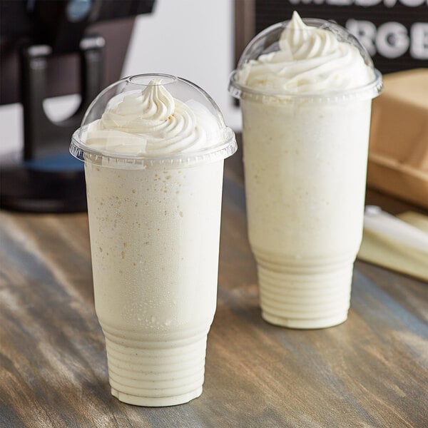 Two Choice clear plastic cups filled with white milkshakes on a table.