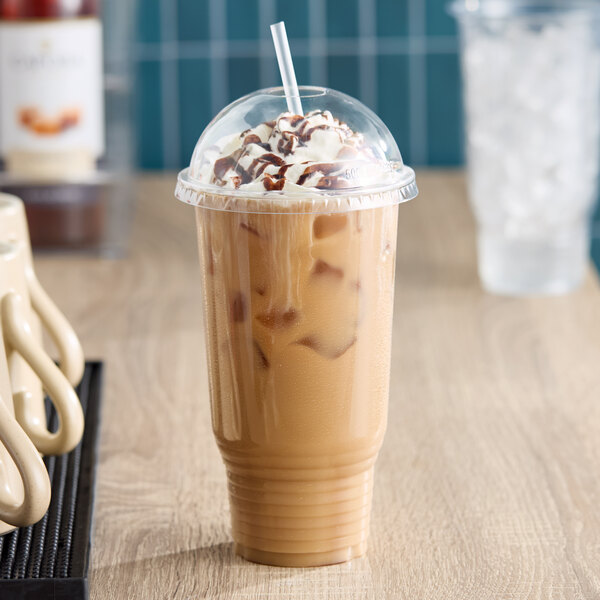 A large clear plastic cold cup with a dome lid, filled with iced coffee topped with whipped cream and chocolate syrup.