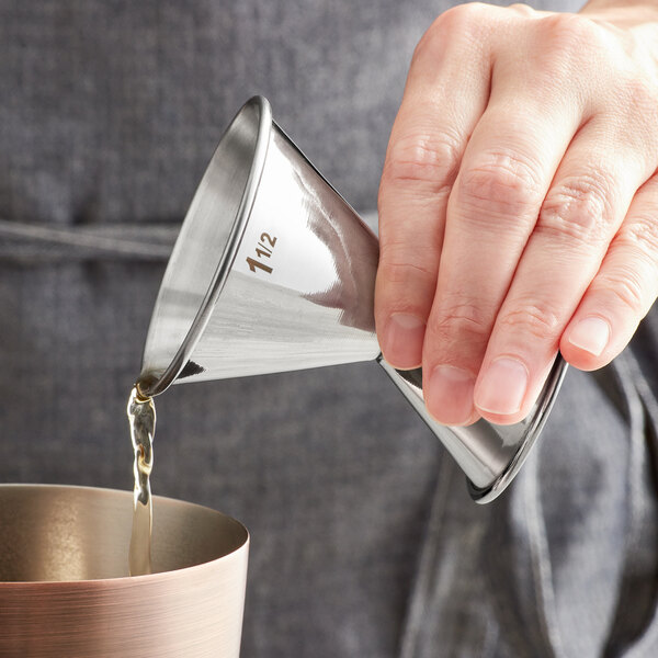 A hand using an American Metalcraft stainless steel jigger to pour liquid into a cup.