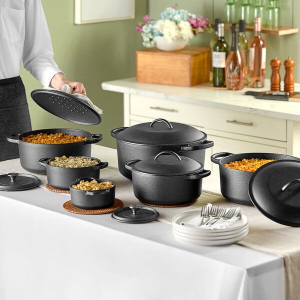 A woman pouring food into a black Valor cast iron Dutch oven on a table with a group of pots and pans.