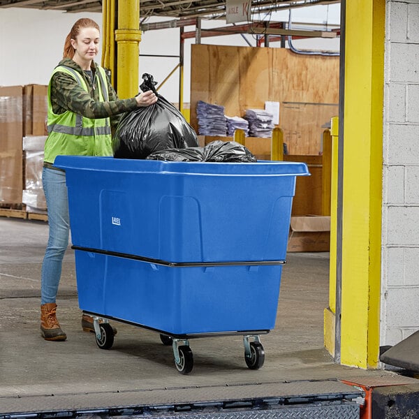 A woman in a green vest pushing a blue Lavex cube truck with a black bag inside.