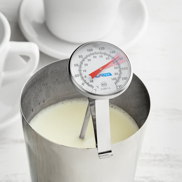 A stainless steel frothing thermometer clipped to a metal pitcher containing milk.