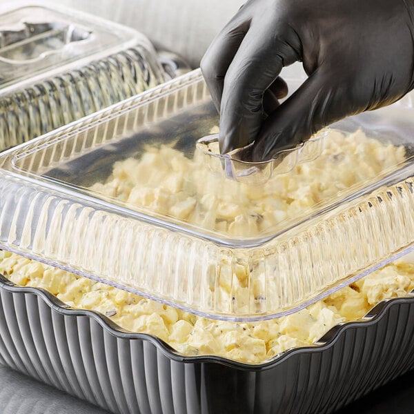 A person wearing black gloves putting food into a black deli crock with clear cover on a counter.