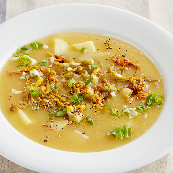 A bowl of soup with Goya zucchini flowers and other vegetables on a white background.