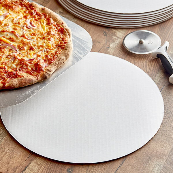 A 16" white corrugated pizza circle with a pizza cutter next to a pizza on a table.