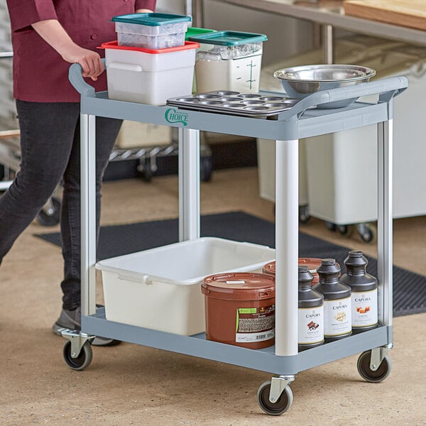 A woman pushing a gray Choice utility cart with two shelves holding food containers.