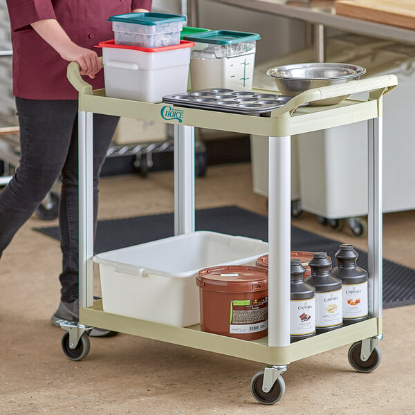 A woman pushing a Choice beige utility cart with food containers on the shelves.