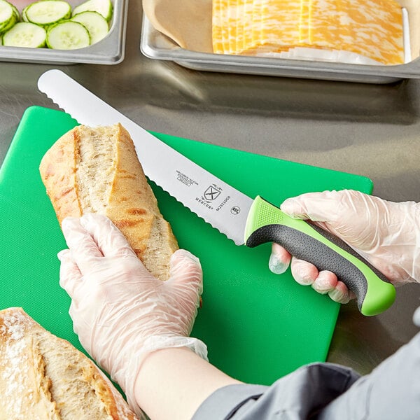 A person's hand holding a Mercer Culinary Millennia Colors bread knife and cutting a loaf of bread.
