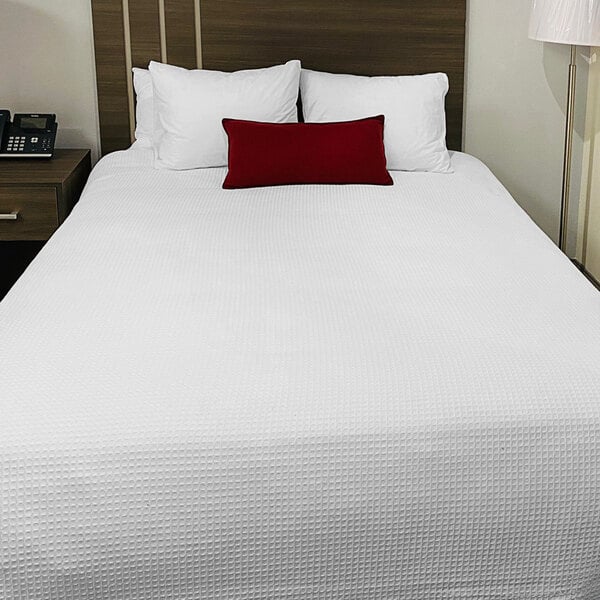 A white bed with a Oxford white Jaipur thermal honeycomb hotel blanket on it and a red pillow.