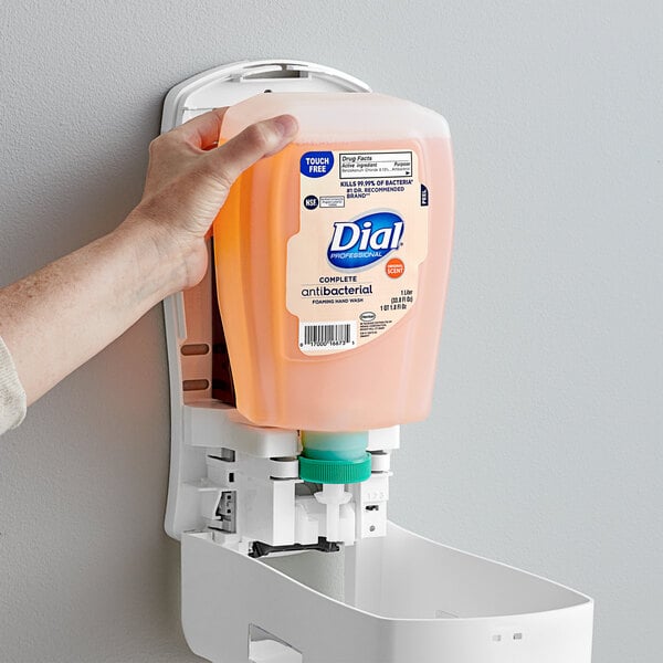 A hand holding a Dial foaming hand soap refill bottle in front of a wall mounted soap dispenser.