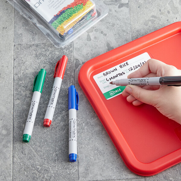 A hand holding a Dri Mark fine tip permanent marker and writing on a red tray.