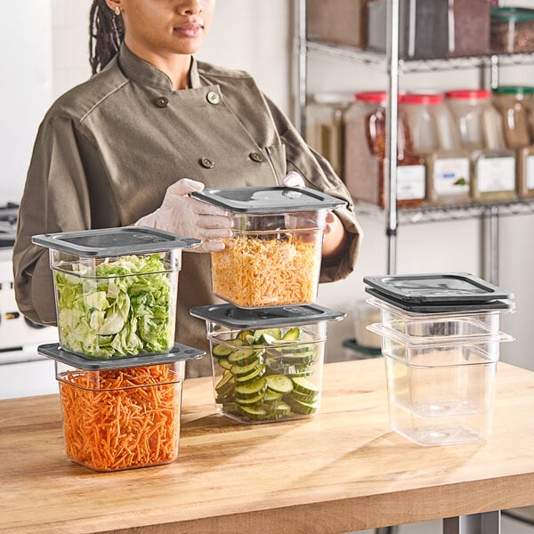 A woman in a kitchen holding a Vigor clear plastic food container with cucumbers and carrots.