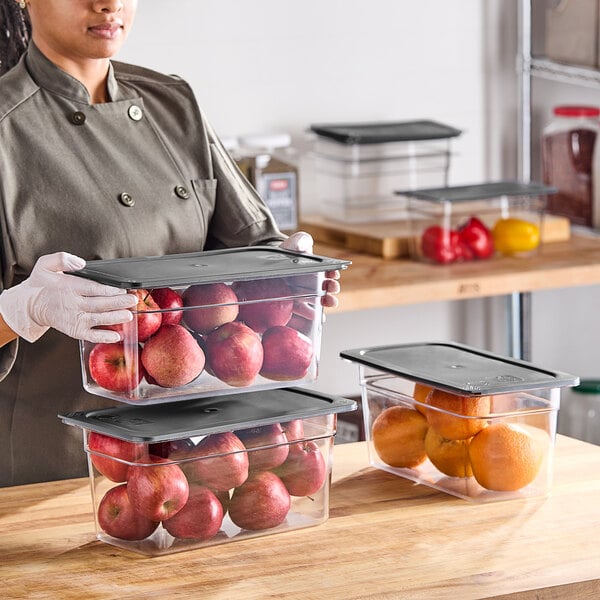 A woman in a chef's uniform holding a Vigor 6-pack of clear plastic food pans with secure sealing covers.