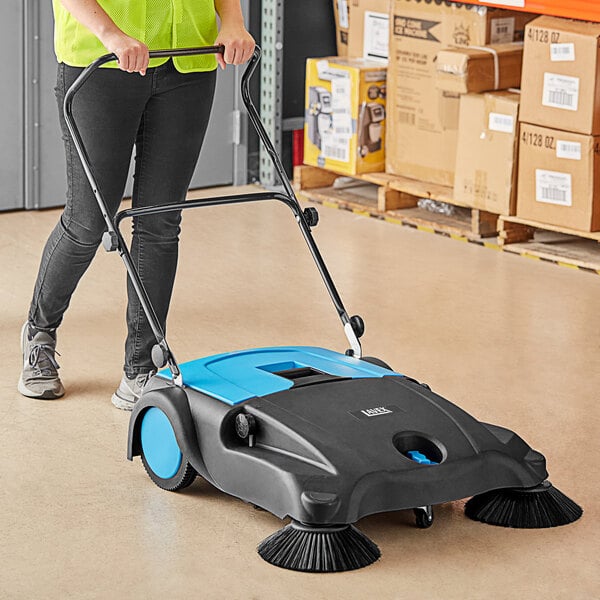 A woman using a Lavex outdoor manual sweeper to clean the floor in a professional kitchen.