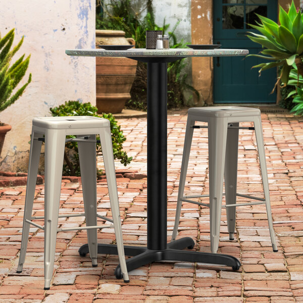A Lancaster Table & Seating Excalibur outdoor table base with a round table surface and white stools on a brick patio.