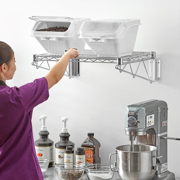 A woman in purple scrubs using a Baker's Lane wall-mounted ingredient bin shelf to store a container of brown food.