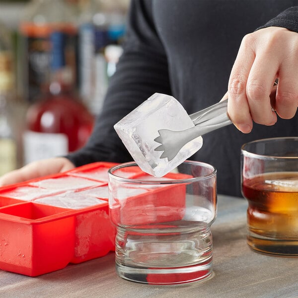 A person using red Choice silicone tongs to put ice cubes in a glass.