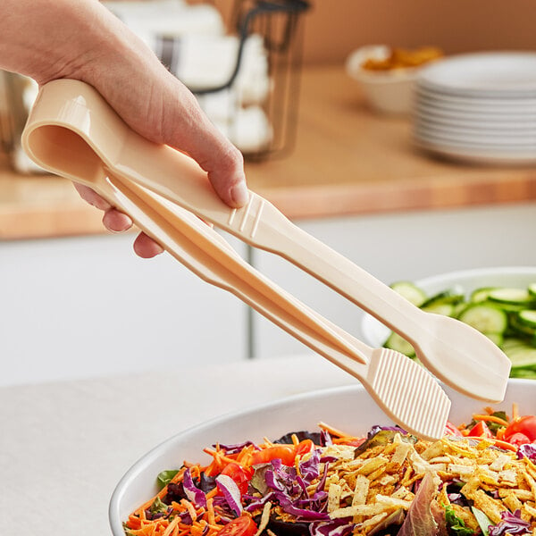 A person holding Thunder Group Beige Polycarbonate Flat Grip Tongs over a bowl of salad.