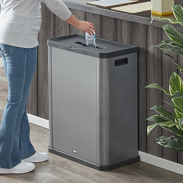 A woman standing next to a Rubbermaid dark gray shell-shaped recycling container with a plastic bag in it.