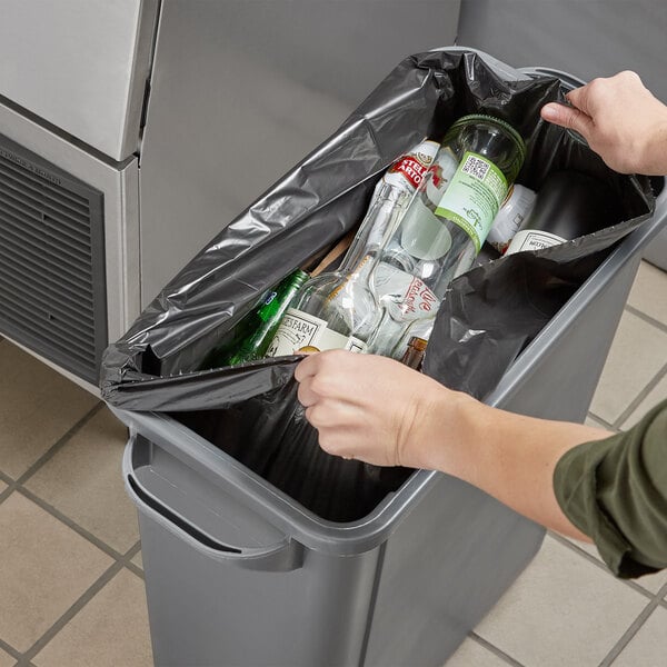 A person putting a Lavex Hercules black trash bag full of bottles into a trash can.