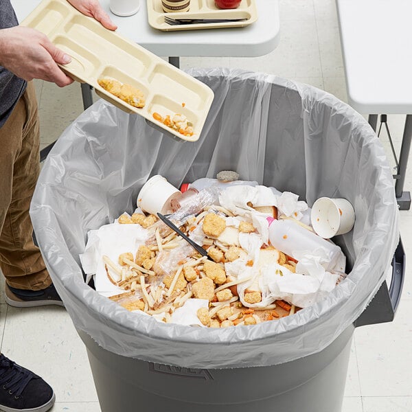 A person holding a tray of food dumping it into a white trash bag in a trash can.