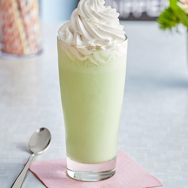 A glass of milkshake with green J. Hungerford Smith Creme De Menthe syrup and whipped cream with a spoon on a napkin.