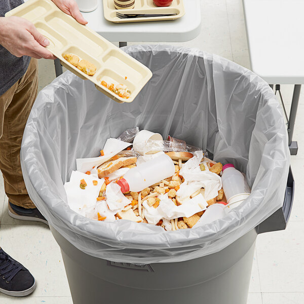 A person holding a tray of food with a fork and knife dumping it into a trash can lined with a Lavex Hercules clear trash bag.
