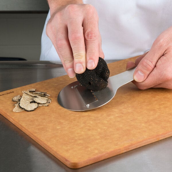 A hand using an Ateco chocolate shaver to peel a mushroom on a cutting board.