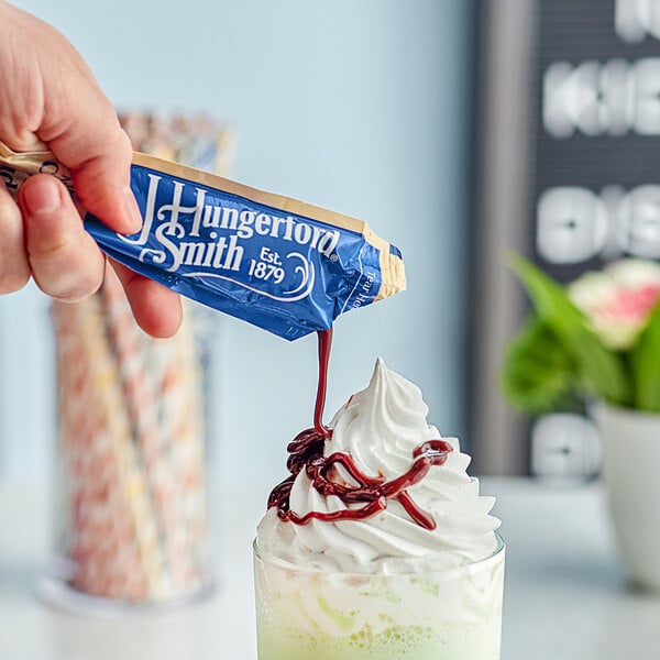 A hand pouring a J. Hungerford Smith Chocolate Fudge portion packet into a drink.