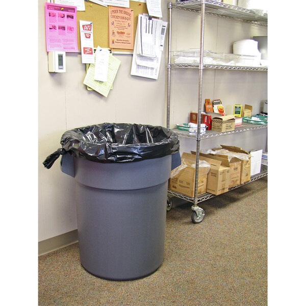 A grey trash can with a black Lavex Hercules can liner sitting next to a shelf in a room.
