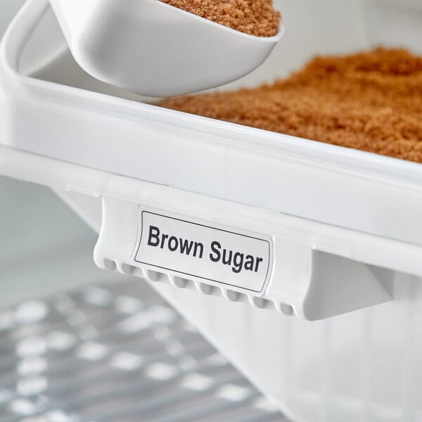 A Baker's Mark ingredient bin label on a container of brown sugar with a scoop of sugar inside.