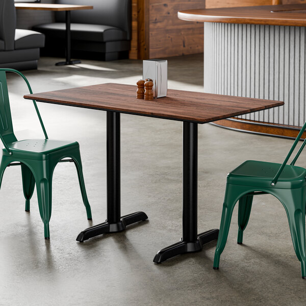 A rectangular standard height table with a textured walnut finish top and two black end base plates, shown with green metal chairs in a restaurant setting.