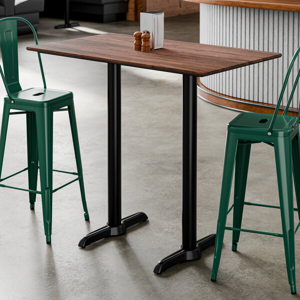 A rectangular bar height table with a textured walnut finish top and two black metal base plates.