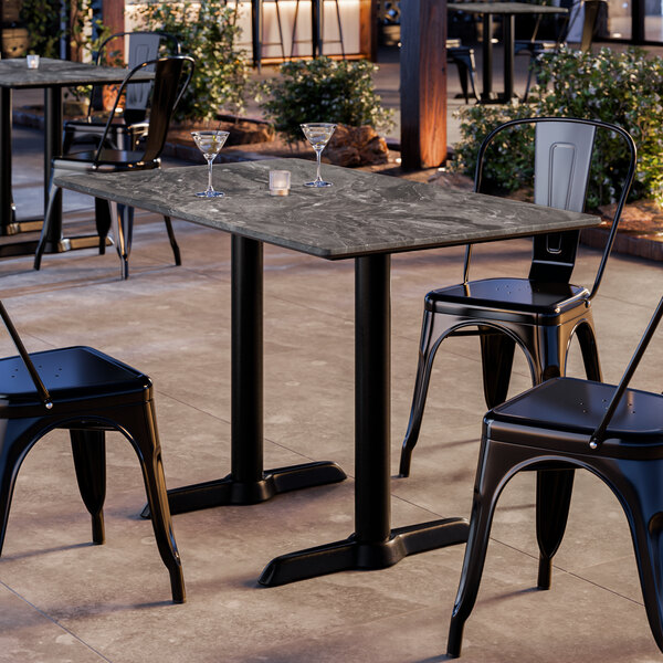 A rectangular outdoor dining table with a smooth dark stone-like finish and two sturdy black metal base plates, surrounded by black metal chairs.