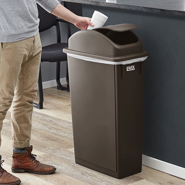 A man standing next to a Lavex brown slim rectangular trash can with a dome swing lid.