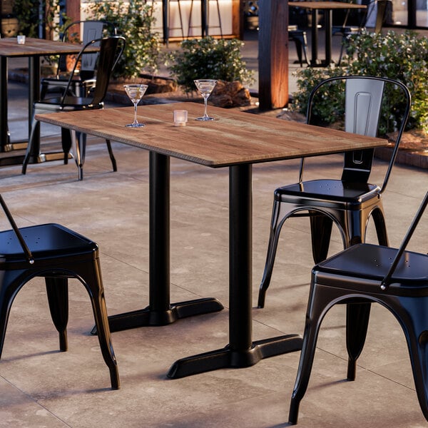 A rectangular standard height table with a textured Yukon oak finish and two black metal end base plates, set outdoors with black metal chairs.