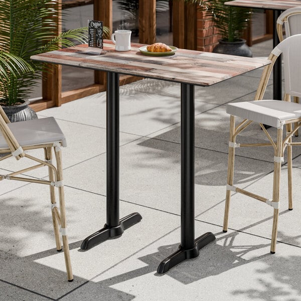 A rectangular bar height table with a textured mixed plank finish and two black metal end base plates, designed for outdoor use.