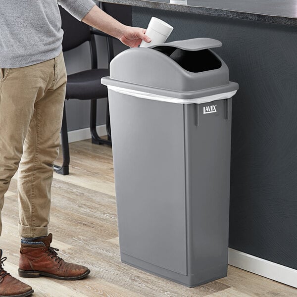 A person standing next to a Lavex gray rectangular trash can with a dome lid.