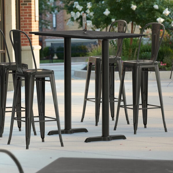 A Lancaster Table & Seating rectangular bar height table with grey surface and white line on an outdoor patio with black bar stools.