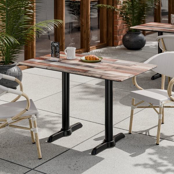 A rectangular standard height table with a textured mixed plank finish and two black end outdoor base plates.