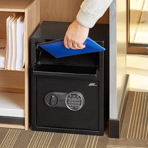 A person opening a black steel depository safe with an electronic keypad lock.