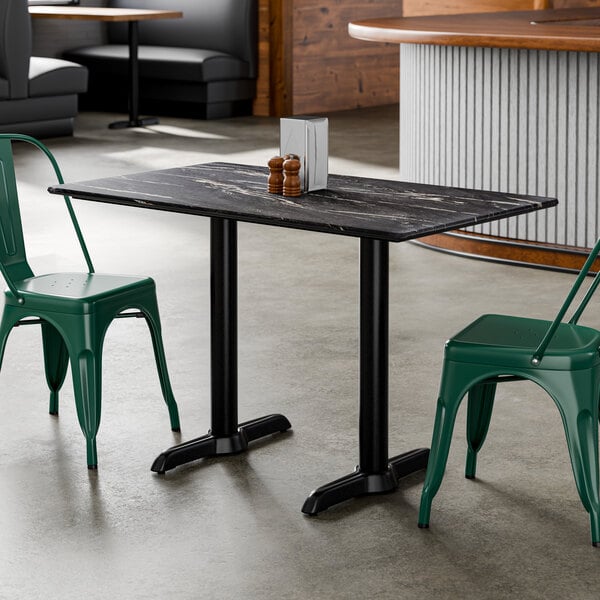 A rectangular standard height table with a smooth dark finish and two black end base plates, shown with green metal chairs in a restaurant setting.