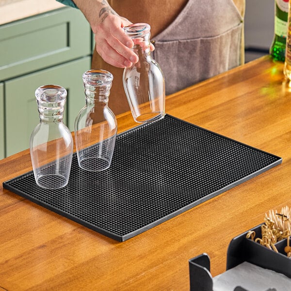 A person pouring drinks into glasses on a black TableCraft bar mat over a table.