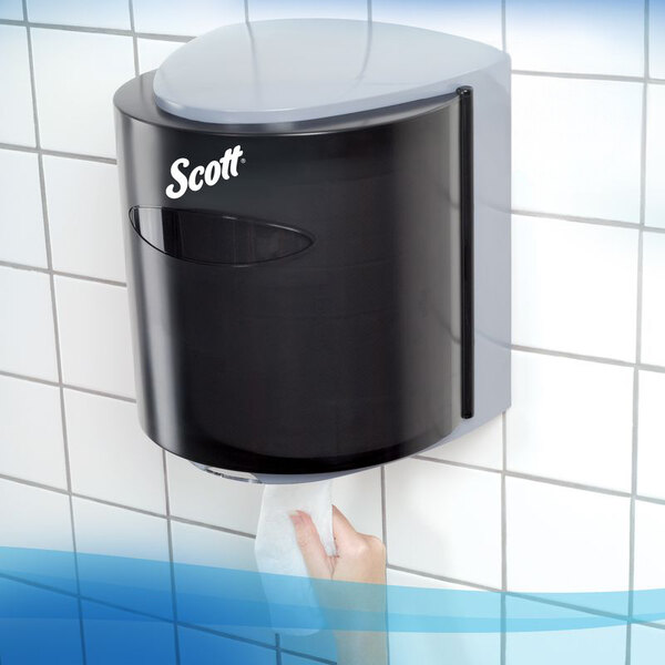 A black wall-mounted paper towel dispenser with a hand pulling a towel from the bottom, installed on a tiled wall.