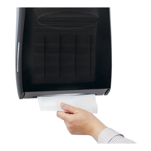 A hand pulling a paper towel from a wall-mounted black dispenser.