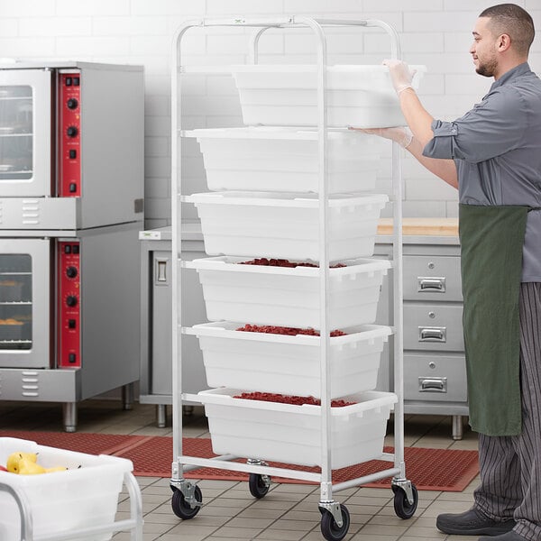 A man in an apron using a Regency mobile aluminum lug rack to move white tote boxes filled with food.