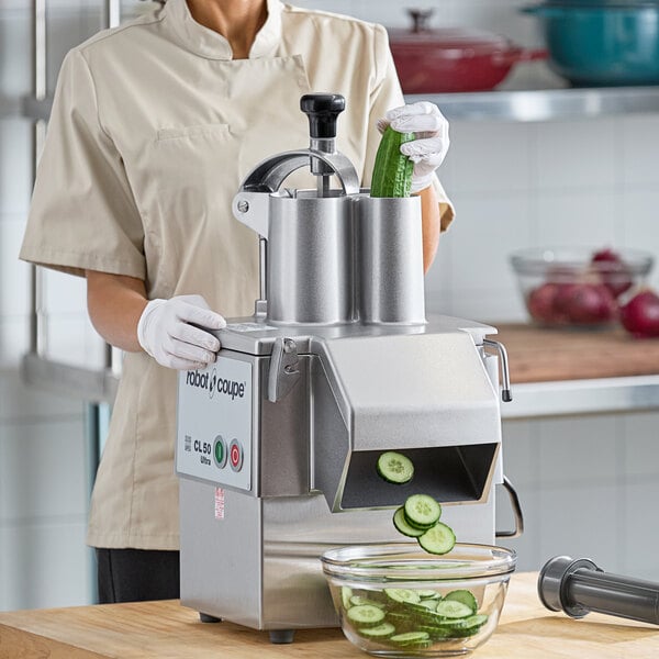 A woman in a white coat and gloves using a Robot Coupe food processor to slice cucumbers in a professional kitchen.